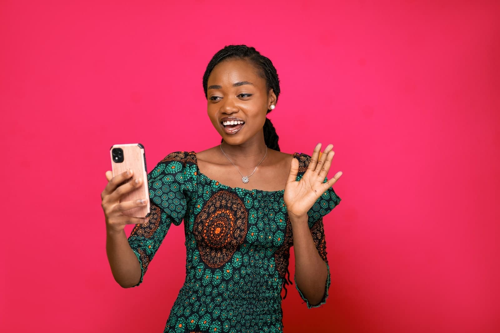 A woman in a green print dress smiles at her phone and waves, against a magenta wall