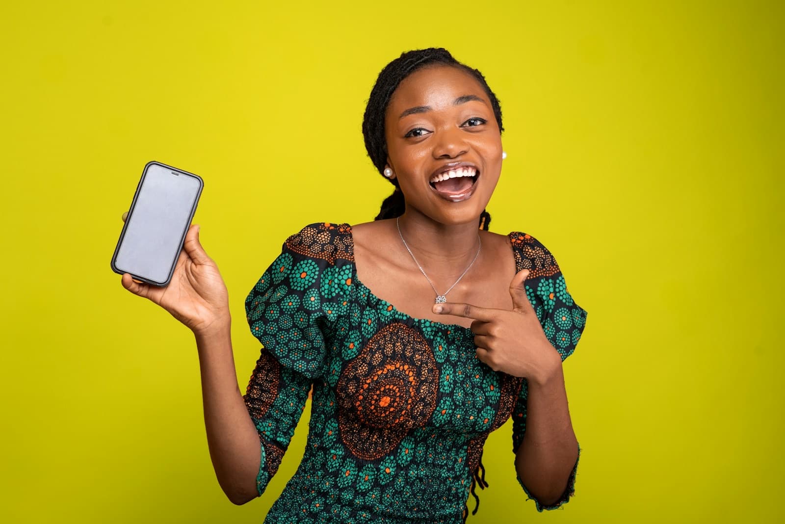 A woman in a green print dress holds up her phone and smiles, against a yellow wall